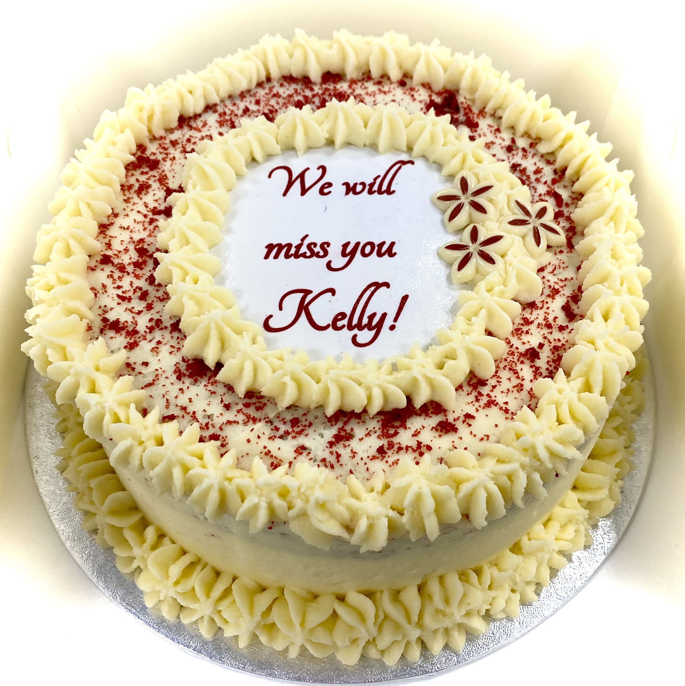 Round Red Velvet Cake with ornate white icing borders and red sprinkles, displaying the message 'We will miss you Kelly!' in the center. Two red and white flower decorations are on the top right side. Perfect for a special occasion, this cake is topped with delicious cream cheese frosting.